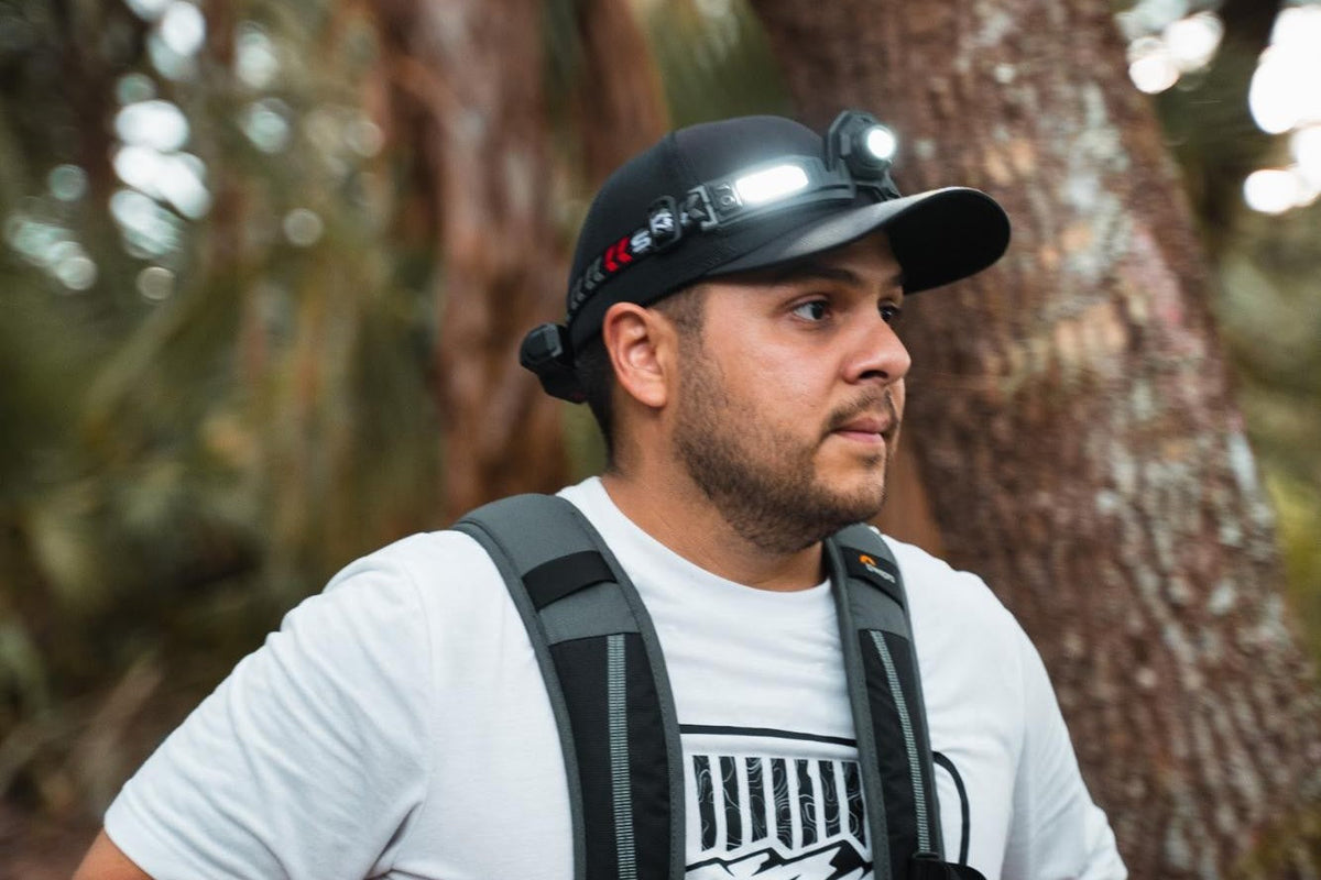 Male wearing a black hat with a FLEXIT Headlamp on it. He's in a woods setting.