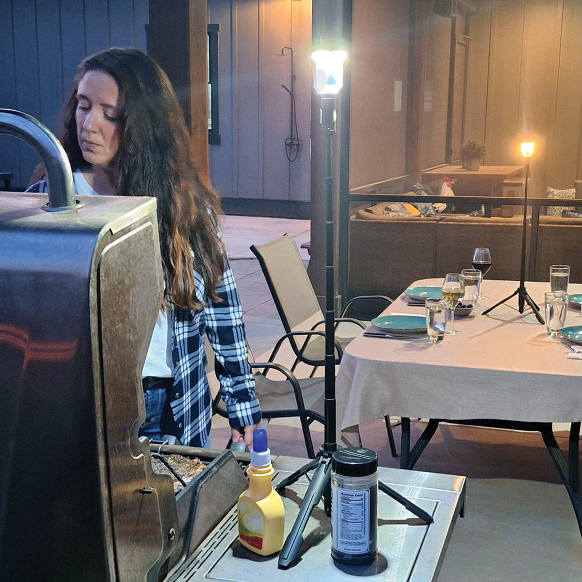 Woman grilling outdoors with a FLi-MORPHE lighting up here grilling experience. In the background, an outdoor table with place settings and wine glasses. A FLi-MORPHE set to warm light sitting in the middle of the table.